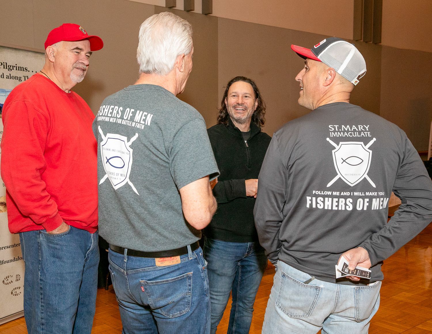 Four people chatting in a room, wearing casual clothes and caps, with two showing matching “Fishers of Men” shirts.