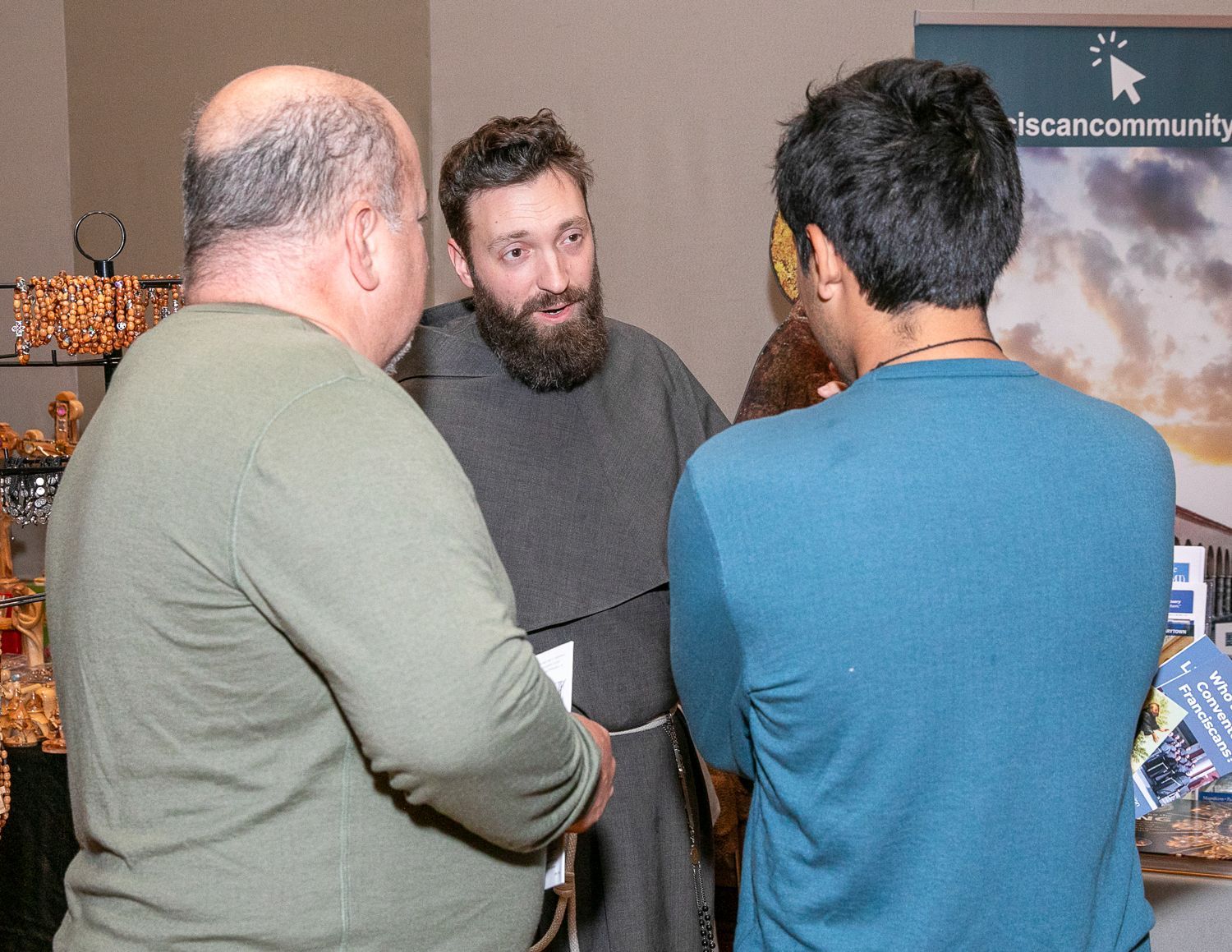 Three people chatting at a booth, with a bearded man speaking to two others indoors.