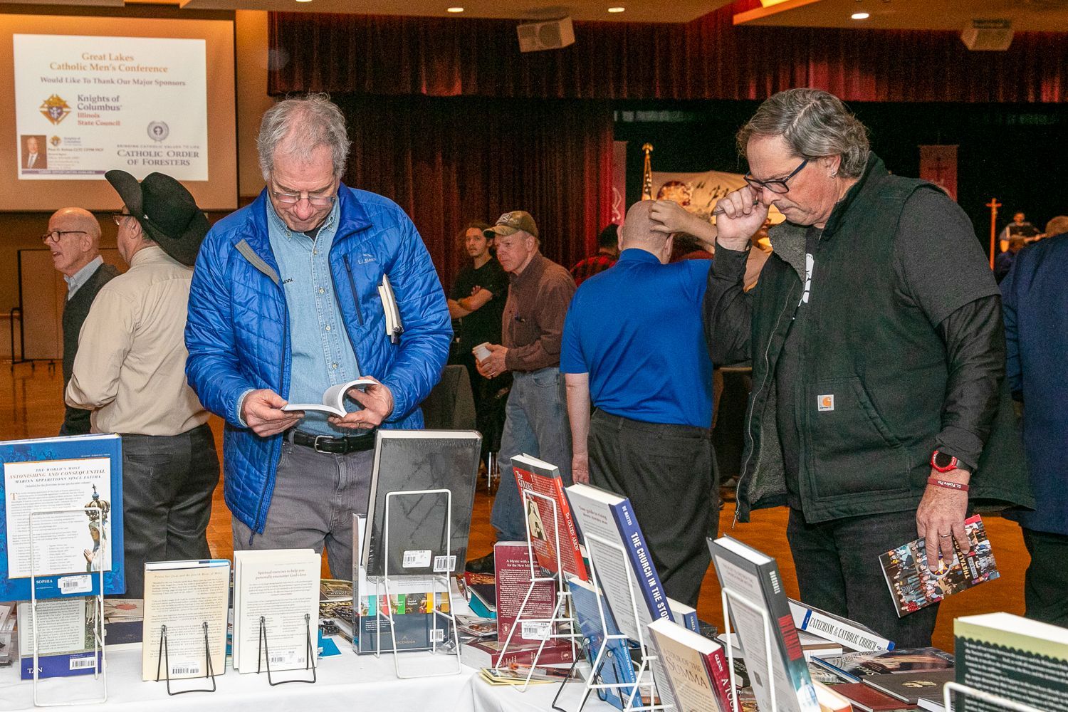 People browsing books and pamphlets at a table in a wood-paneled event hall