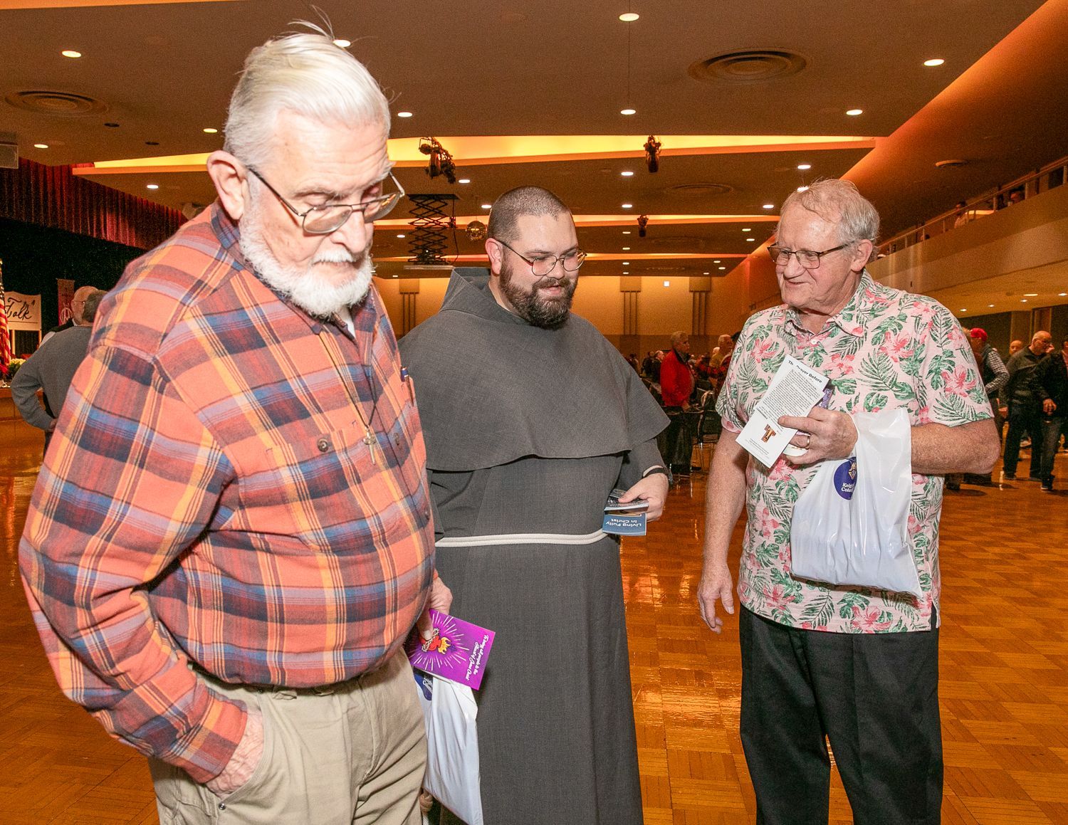 Three men talking in a warmly lit hall, one holding a white cloth and another in a plaid shirt looking down.