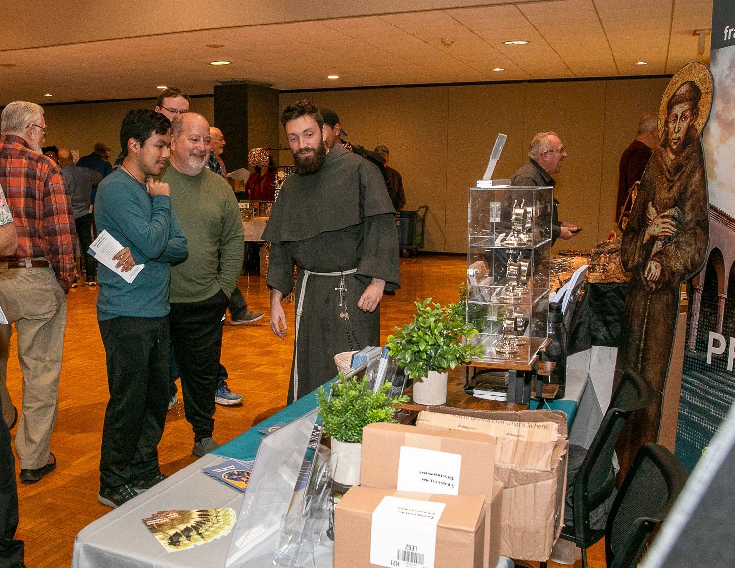 People browsing a booth display at an indoor event, with plants, flyers, and a trophy on the table.