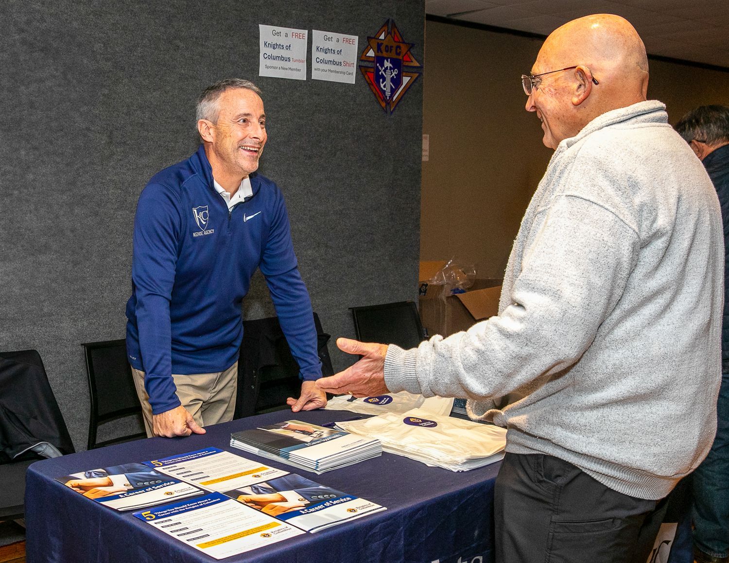 Two men talking across a table at a booth with brochures and papers.