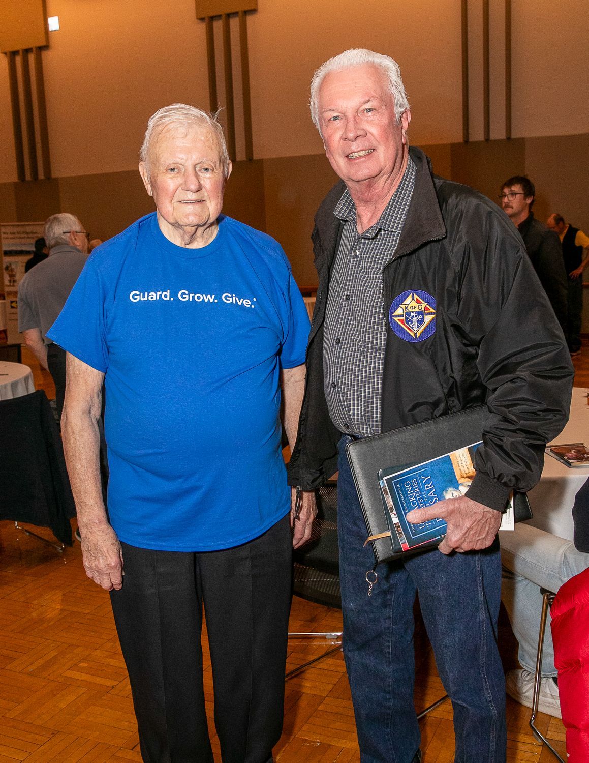 Two people posing indoors, one in a blue shirt and one holding a book and award plaque.