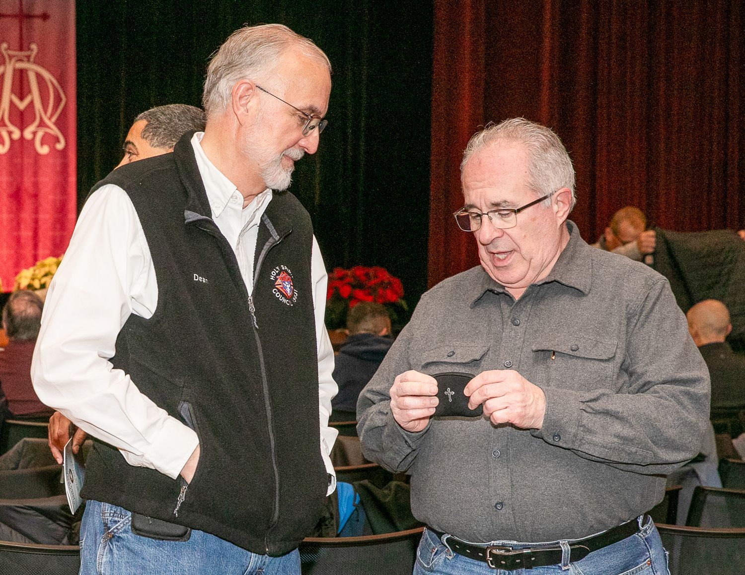 Two men chatting at an indoor event, one holding a small object and the other listening in a black vest.