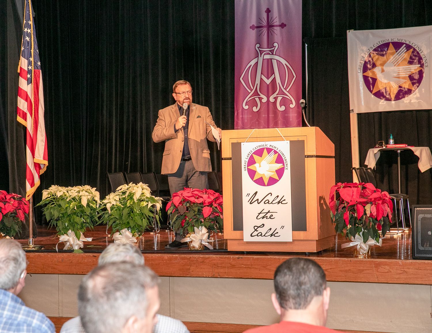 Wayne Messmer at podium on stage with flags, flowers, and a seated audience in front