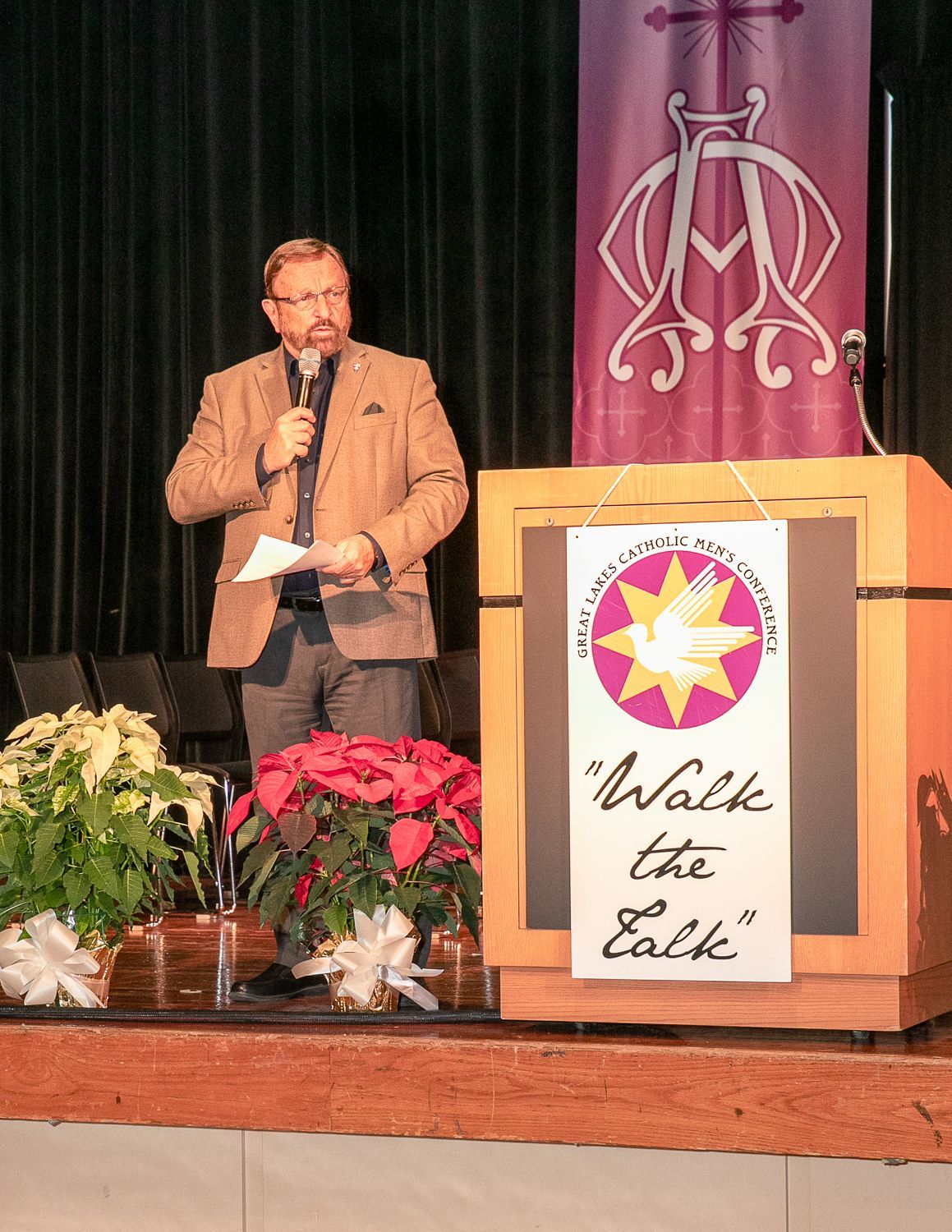 Wayne Messmer at podium on stage beside purple banner and potted flowers, holding a microphone.