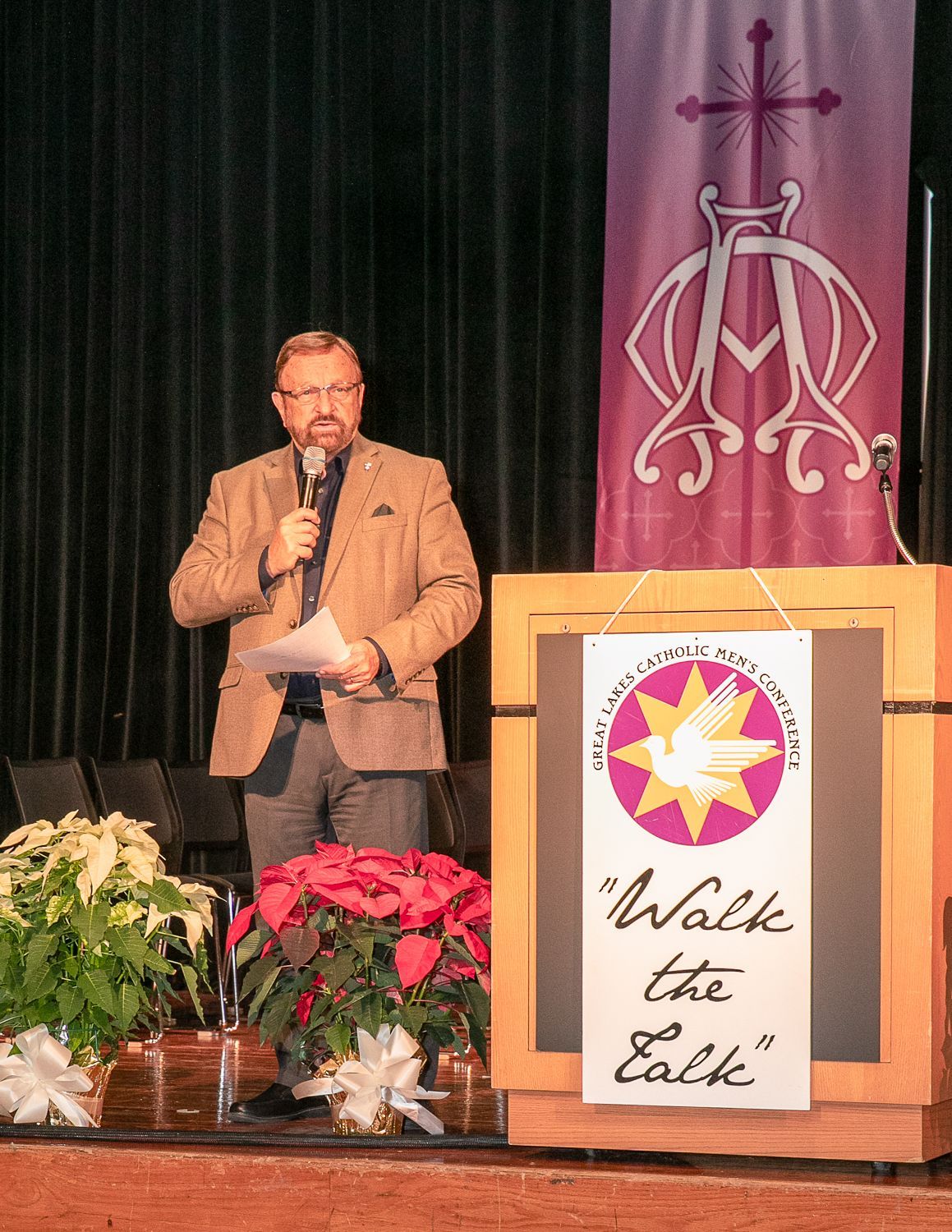 Wayne Messmer speaking at a podium on stage beside a purple banner and a sign reading “Walk the Earth”