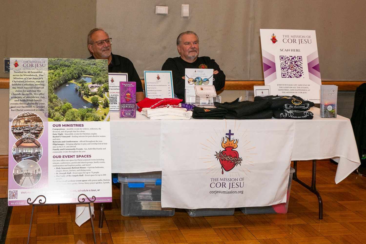 Two men at a vendor table with books, posters, and printed materials at an indoor event