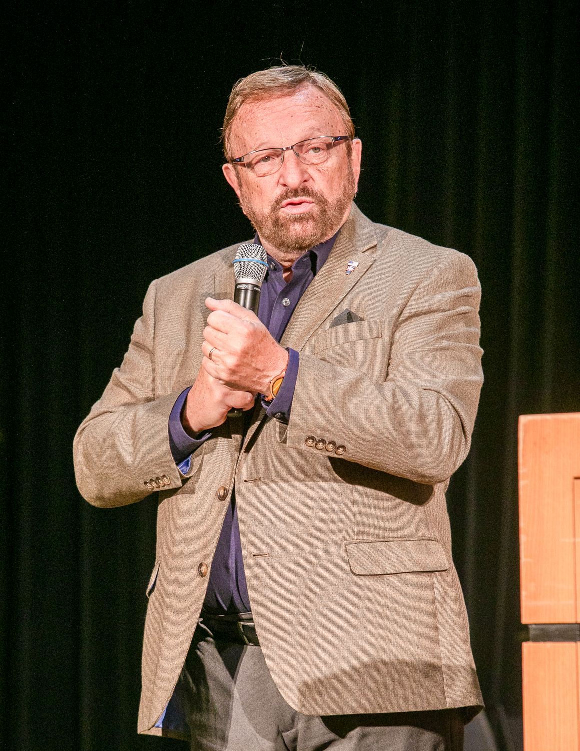 Wayne Messmer, in a tan blazer, speaking into a microphone on a stage with a dark curtain backdrop.