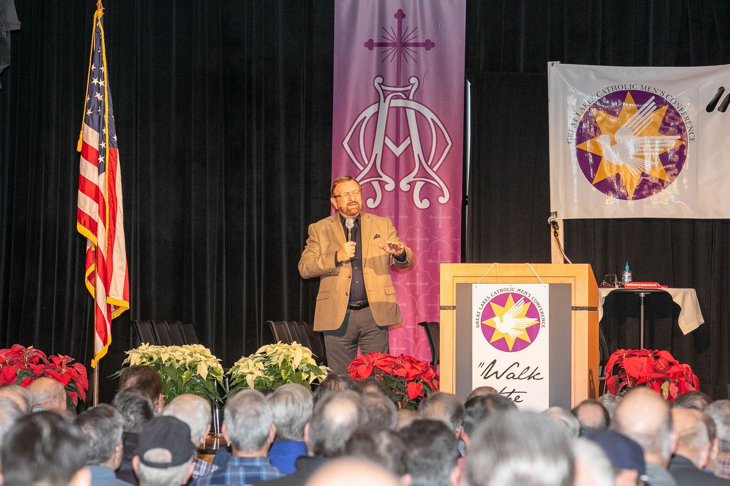 Speaker on stage at podium with flags and banners, addressing audience in auditorium