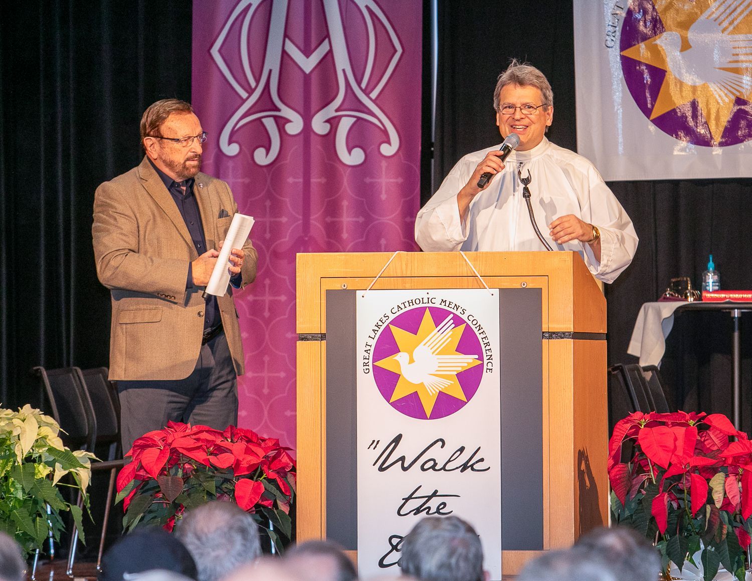 Wayne Messmer and Fr. Max Striedl on stage at a podium with a purple backdrop and poinsettias