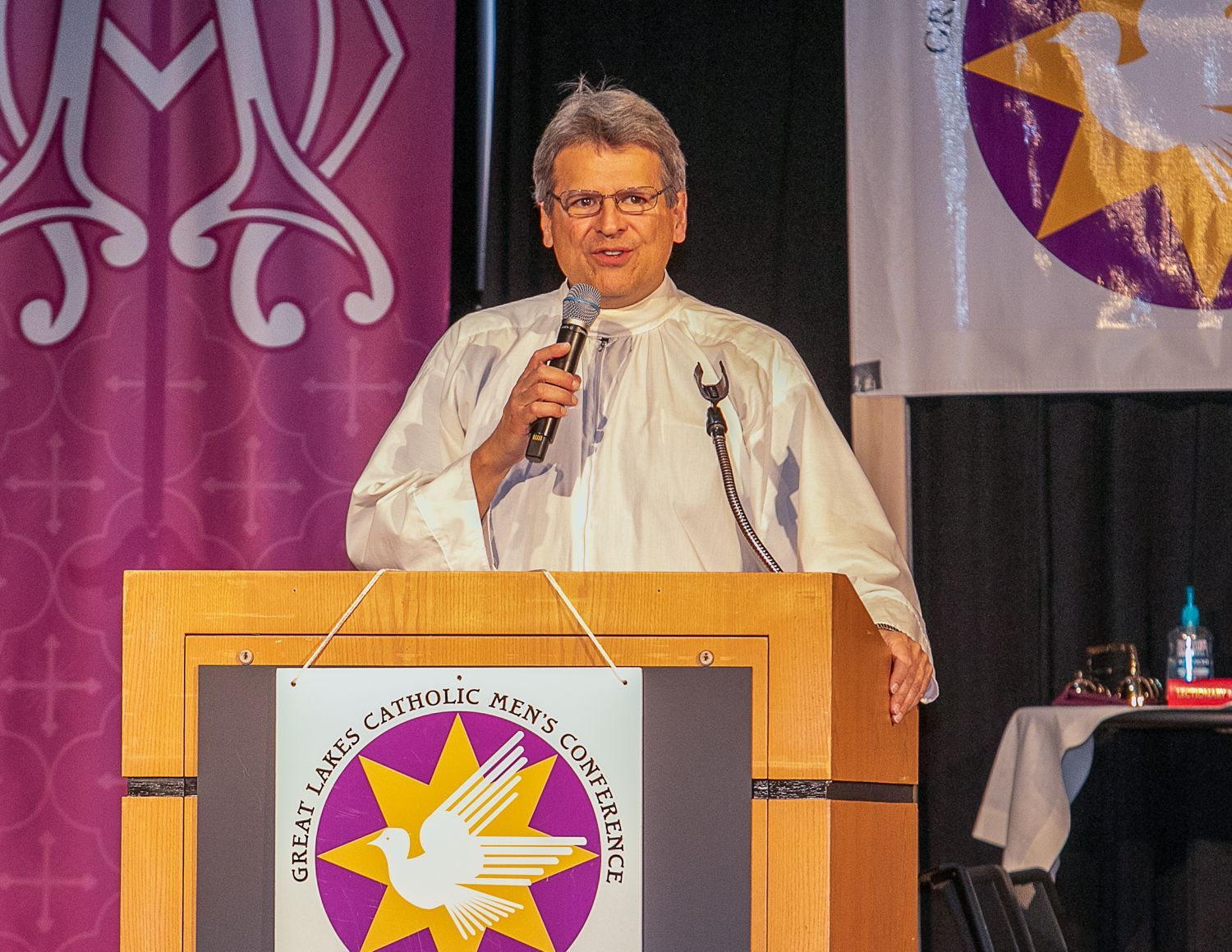 Fr. Max Striedl in a white shirt stands at a podium, speaking into a microphone at a formal event.