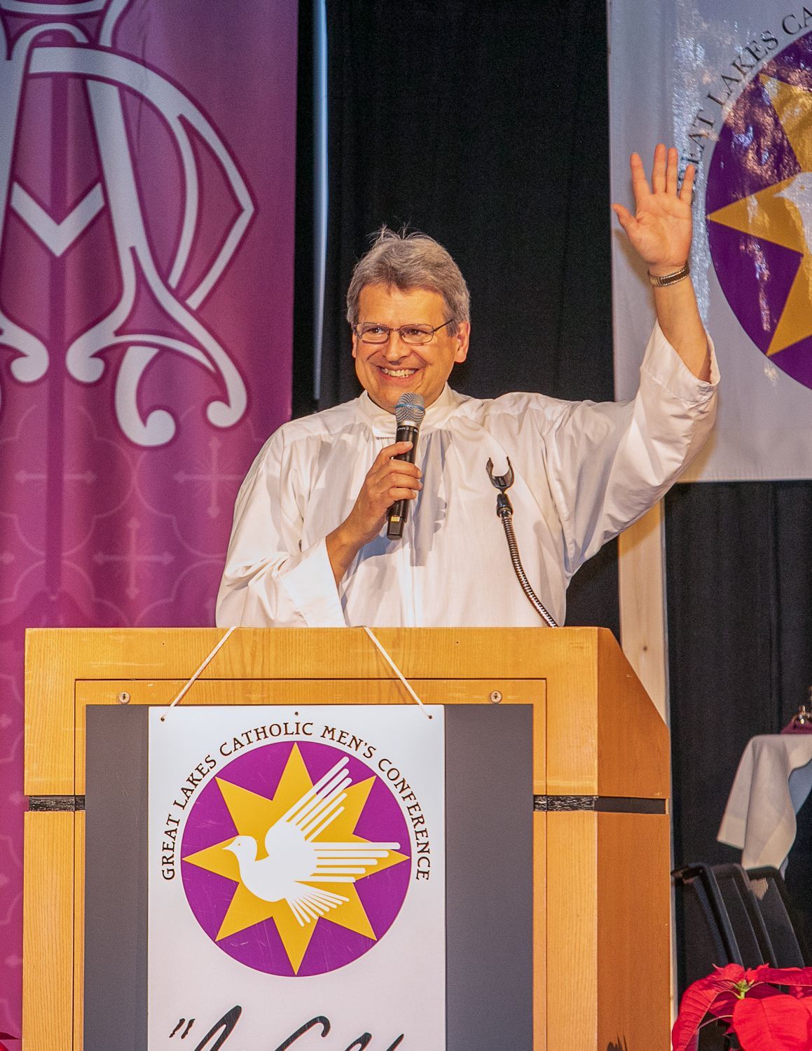 Fr. Max Striedl speaking at a podium, smiling and waving, with purple banners in the background.