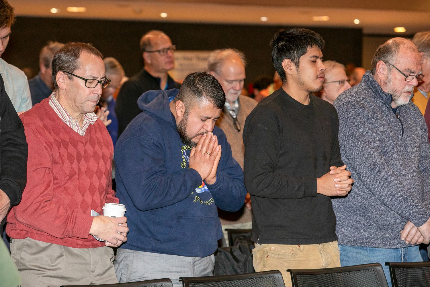 People standing indoors, several with heads bowed and hands clasped in prayer.