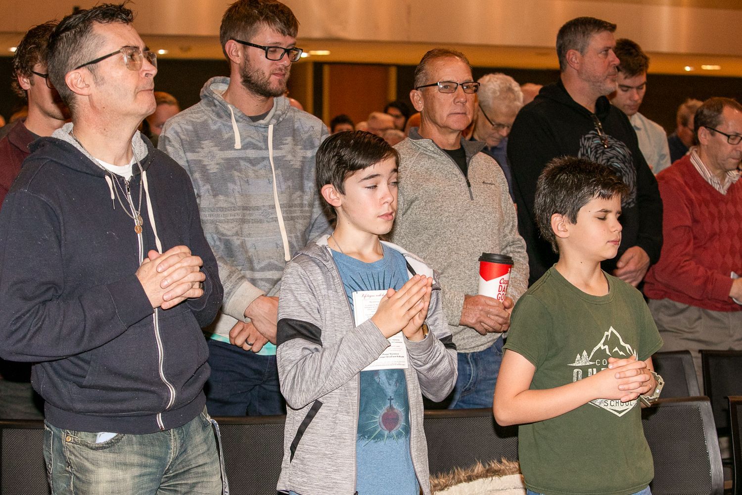 Group of people standing indoors, watching attentively, some holding drinks and cups.