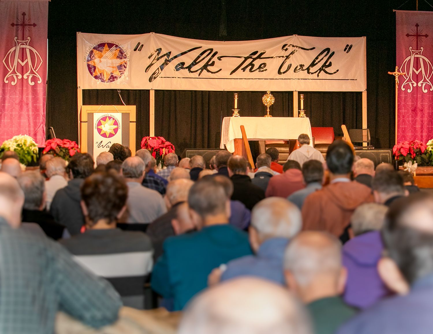 Audience seated in a hall facing a stage with a “Walk the Talk” banner and floral decorations