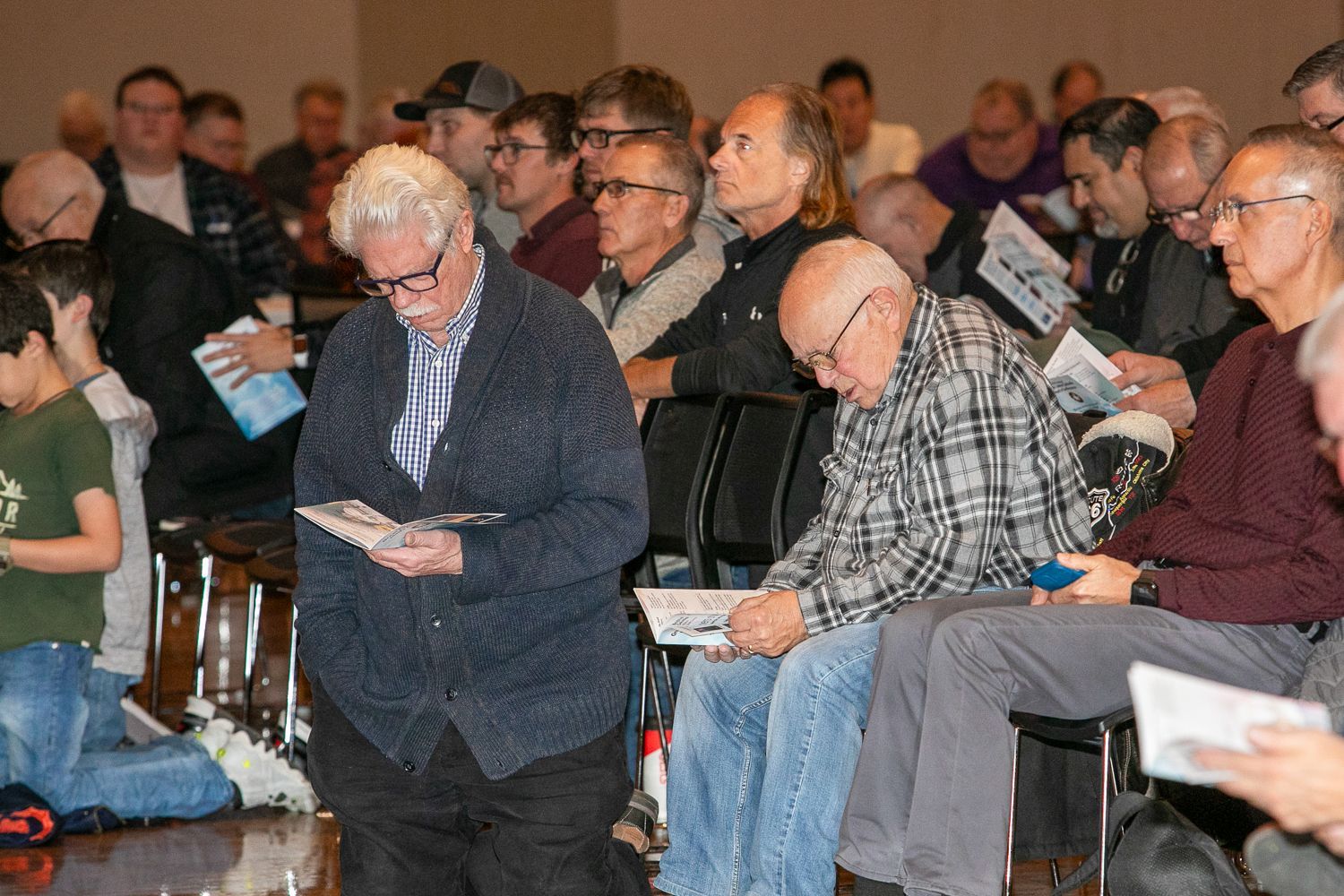 Audience seated indoors, reading programs and waiting during a meeting or event.