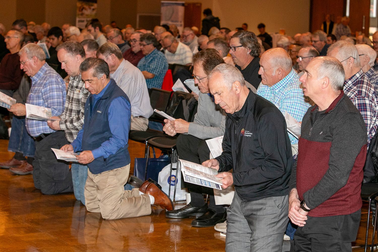 People kneel in a church service, heads bowed in prayer, with wood-paneled walls and chairs behind them.
