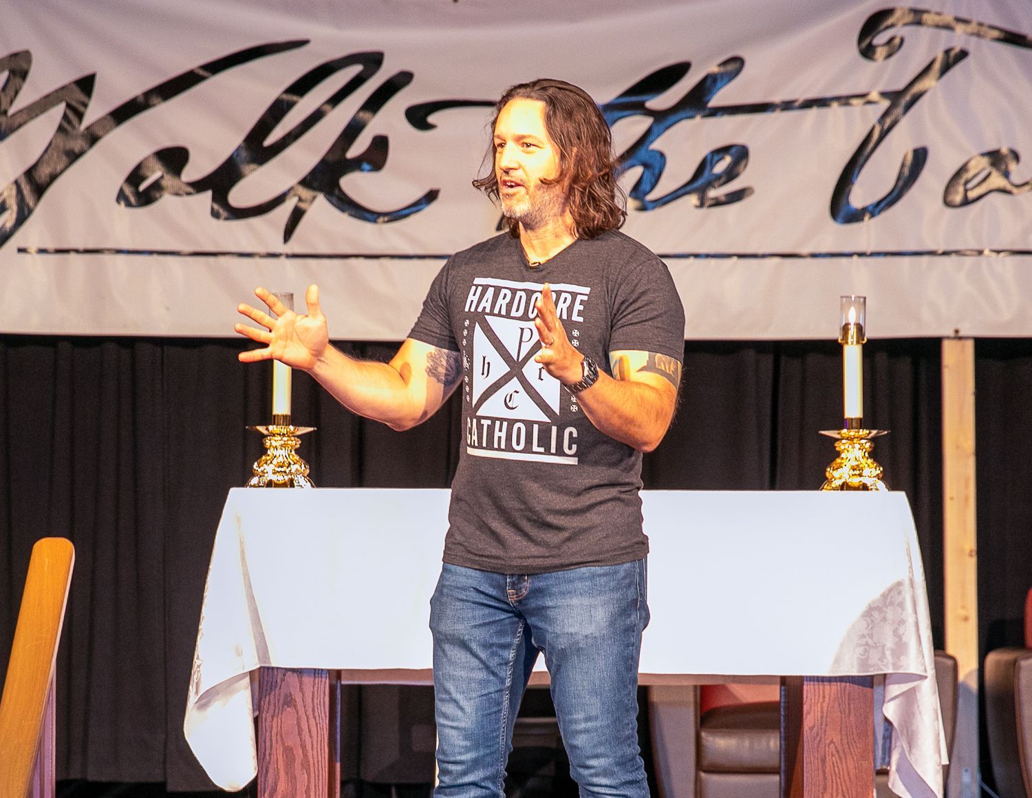 Keith Nester with microphone on stage in front of a table and banner backdrop.