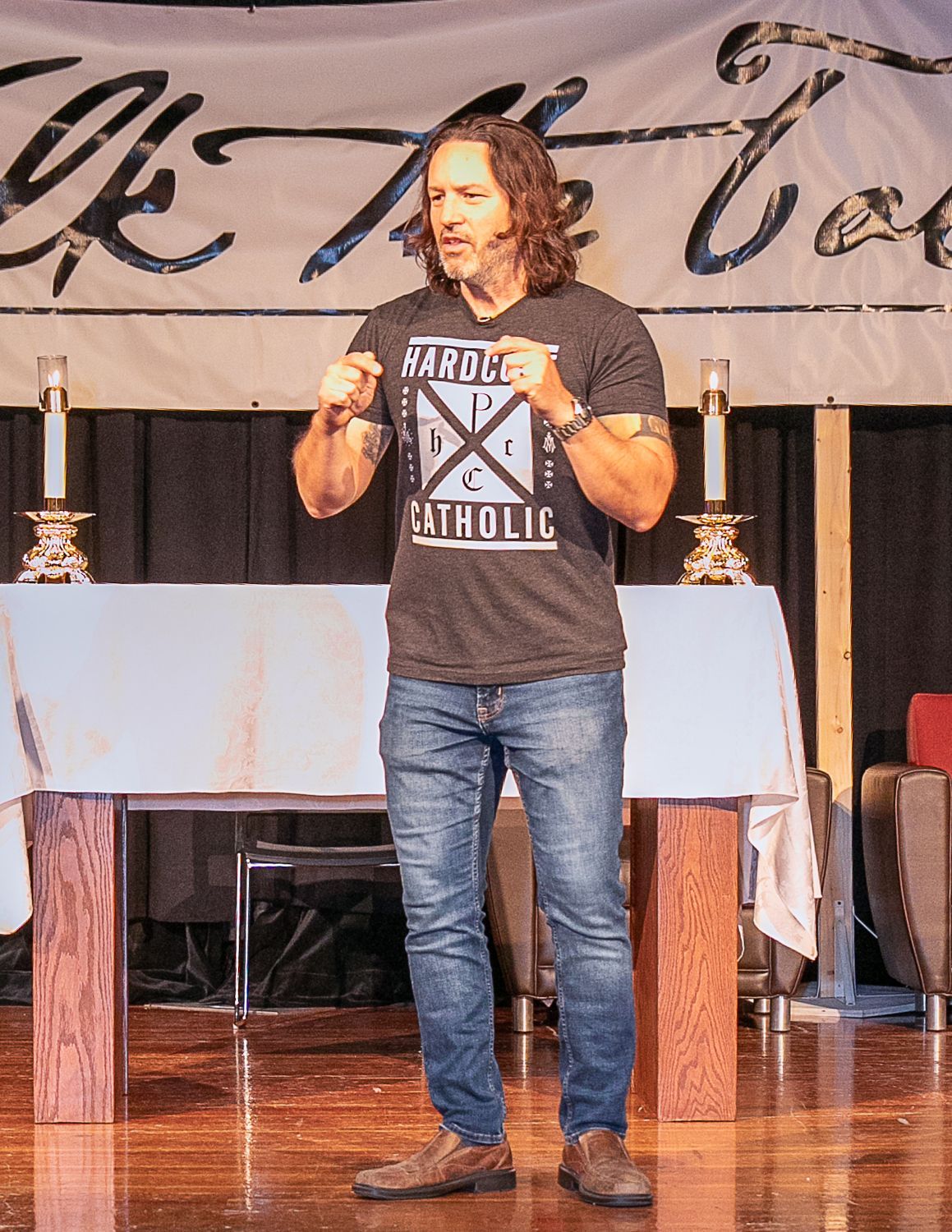 Keith Nester speaking onstage in a black T-shirt, holding a microphone before a table with candles and a banner backdrop