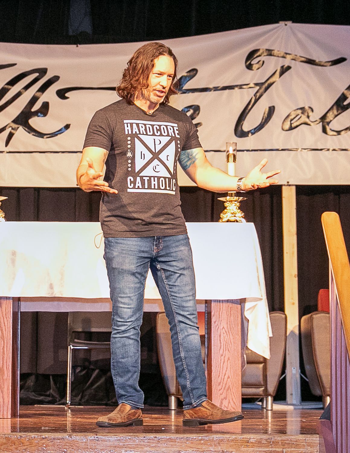 Keith Nester speaking on stage beside a lit candle, wearing a black T-shirt and jeans, in front of a banner