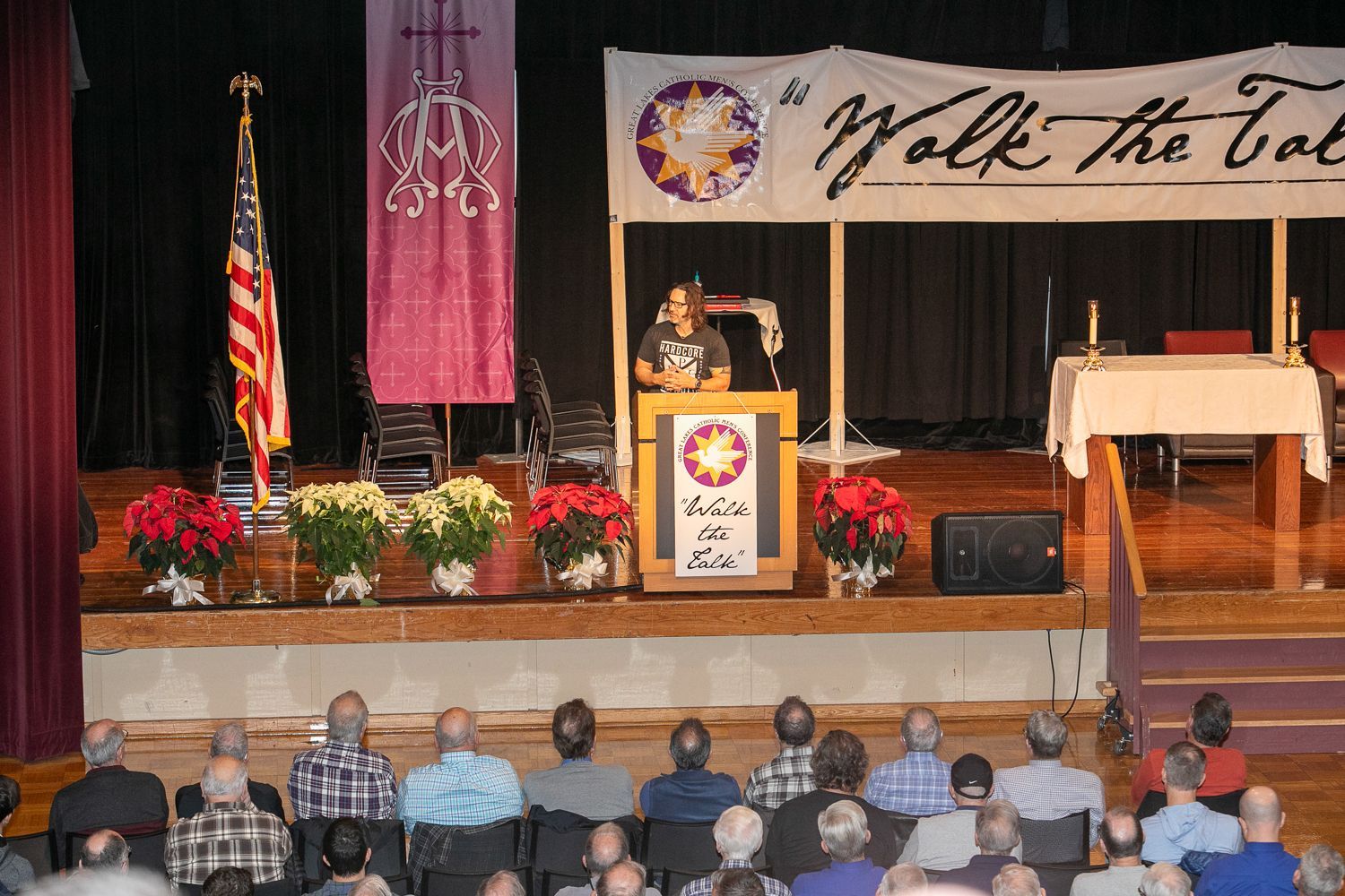 Keith Nester at podium on a stage with a banner, flowers, and seated audience in a hall