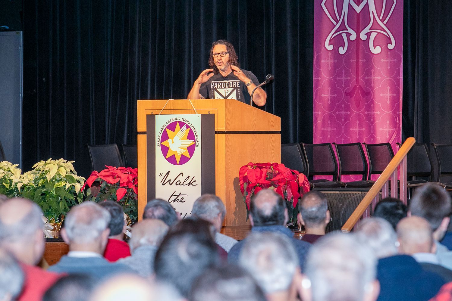 Keith Nester at a podium addressing an audience in a hall, with flowers and banners on stage.