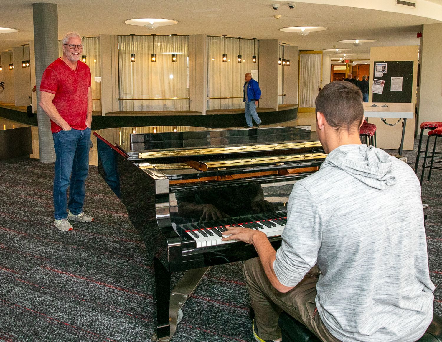 Two people play grand pianos in a lobby, with Sean Forrest standing nearby in a red shirt.