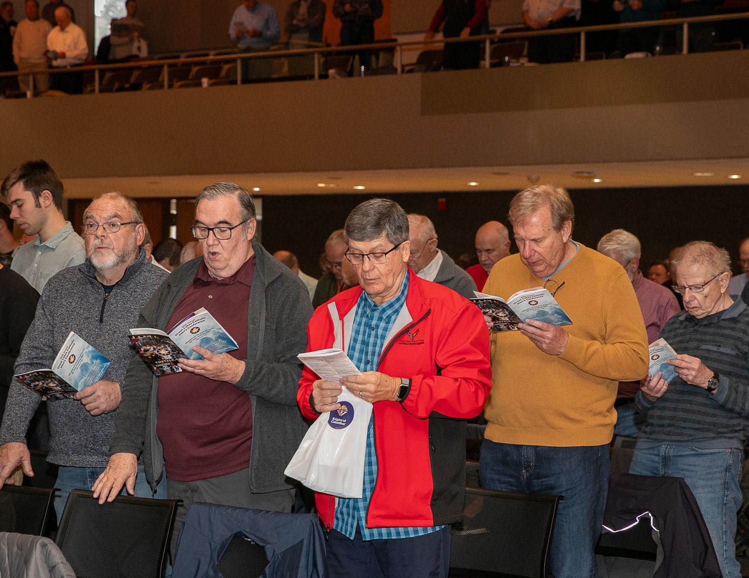 Crowd in a dim auditorium reading pamphlets, including a man in a red jacket and others in casual clothes.