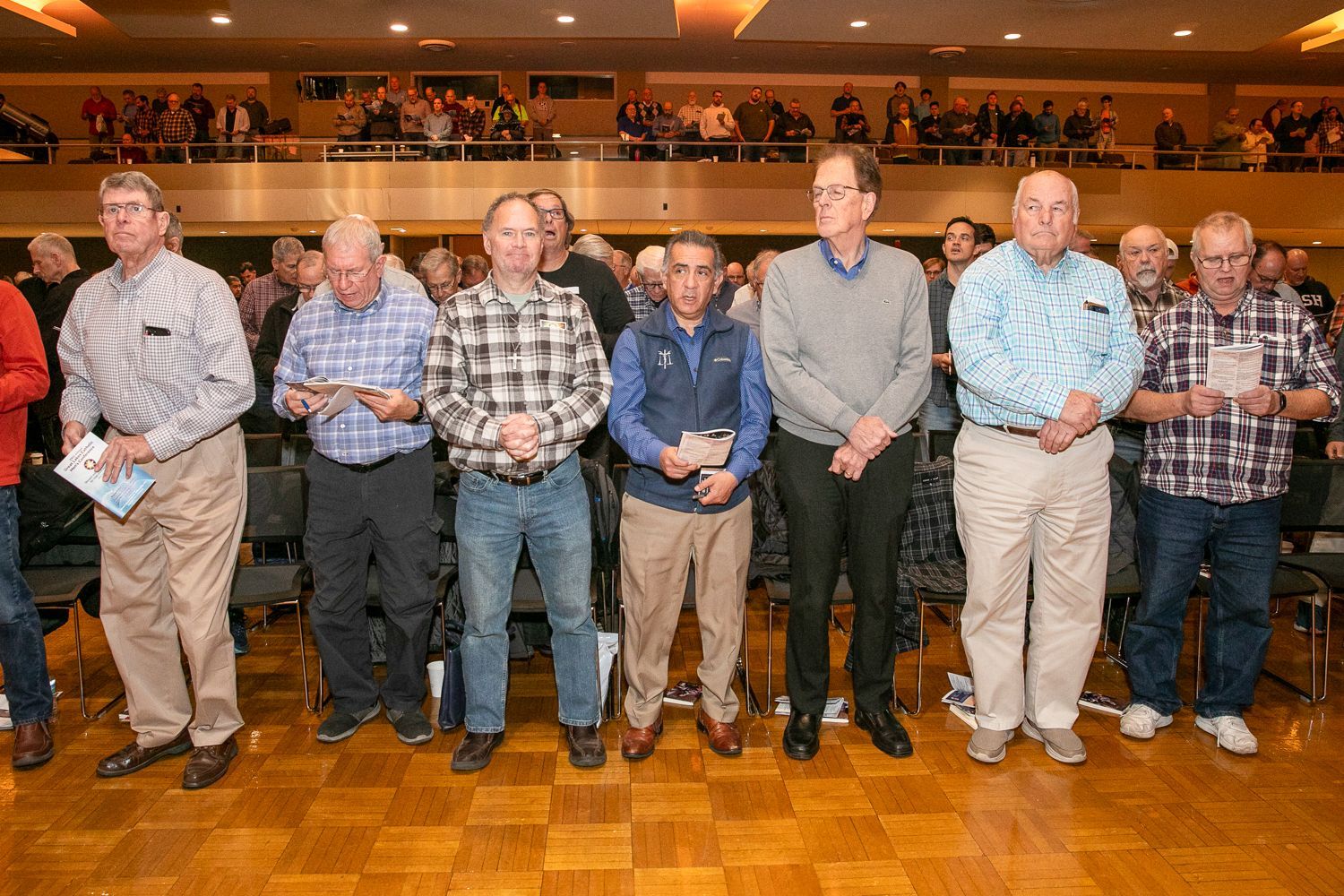 Eight men standing in a row in a banquet hall with warm lighting and wooden floor