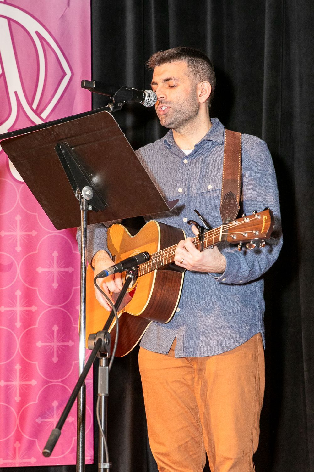 Man playing acoustic guitar and singing into a microphone onstage behind a music stand
