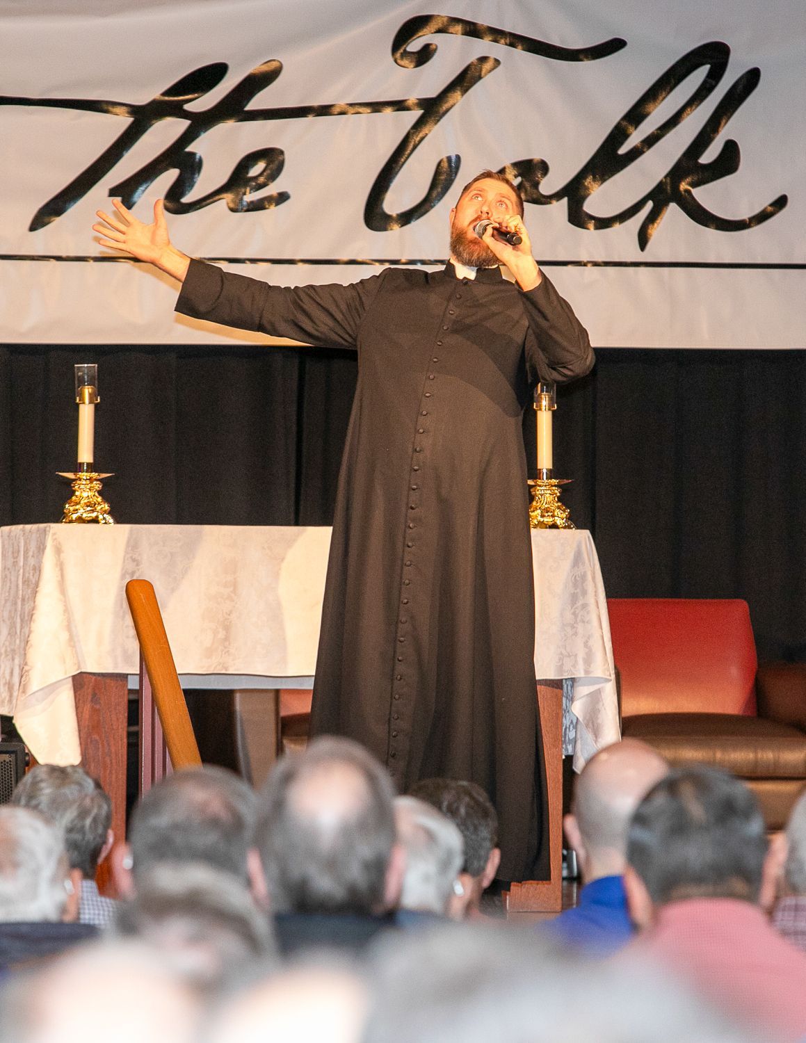 Fr. Sean Grismer in black robe gesturing onstage before a crowd, with “the Talk” banner behind them.