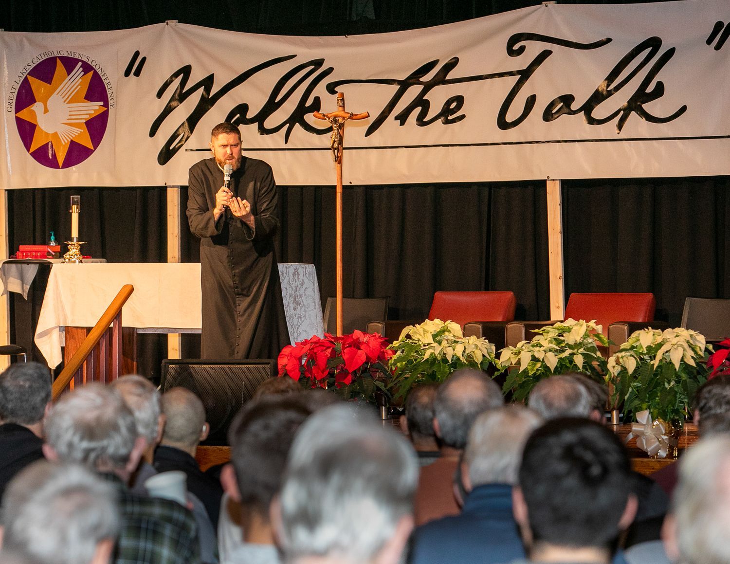 Speaker on stage addressing seated audience beneath a “Walk the Talk” banner