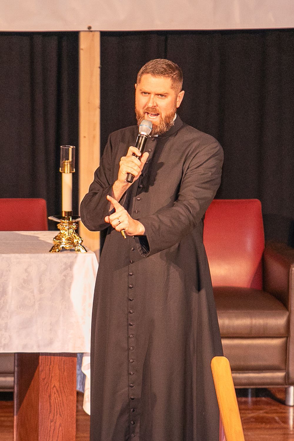 Fr. Sean Grismer speaking into a microphone on a stage beside a table with a candle and red chairs.