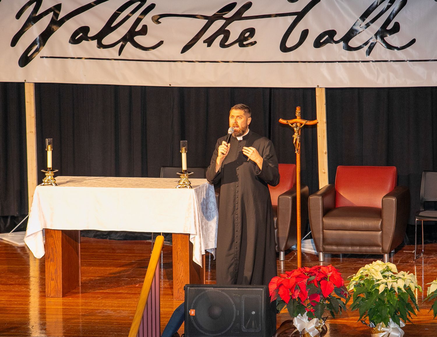Priest speaking on a church stage beside an altar, cross, and red chair, with “Walk the Talk” banner overhead