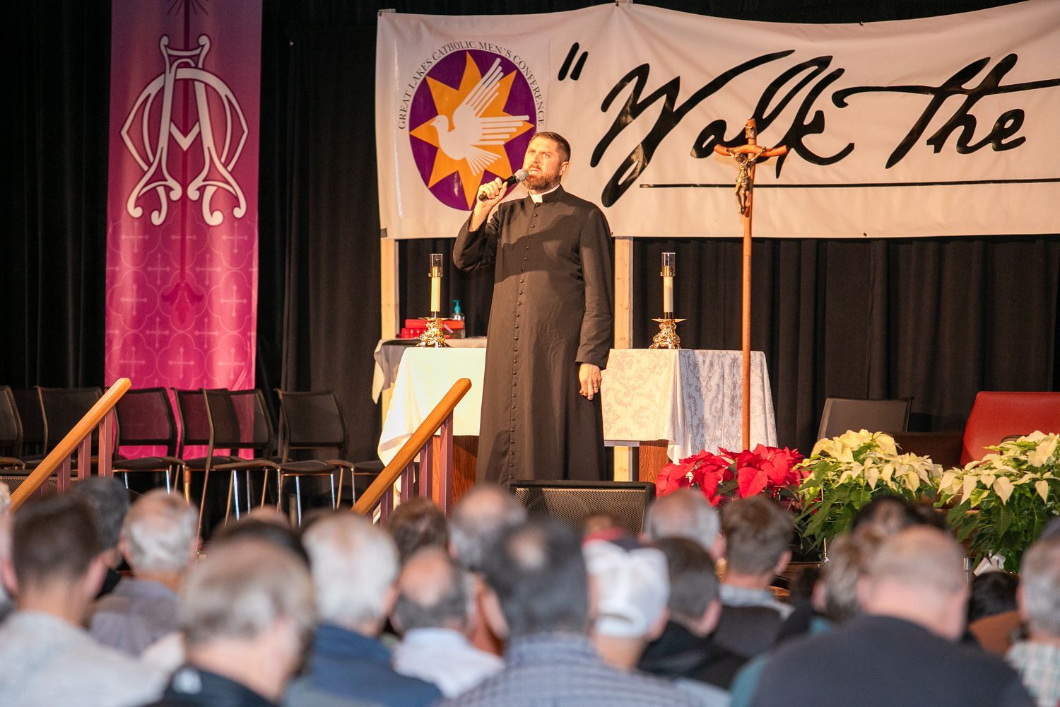 Speaker on stage at a formal event, addressing an audience before a banner and floral arrangement