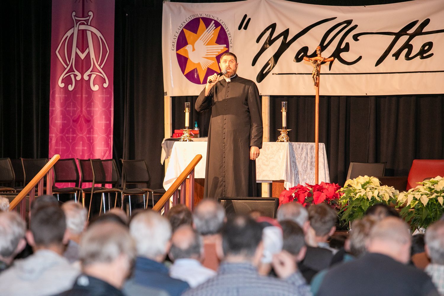 Speaker on stage addressing an audience at a conference, with banners and floral arrangements behind him.