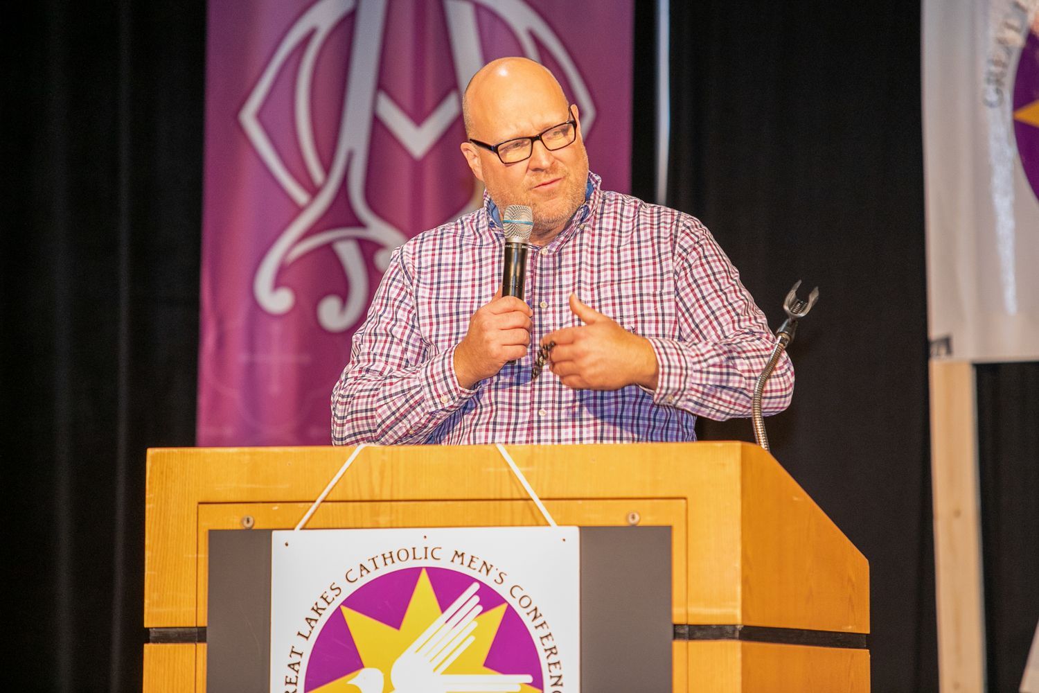Mitch Striedl speaking at a podium on stage, wearing a red plaid shirt, with a purple banner behind him