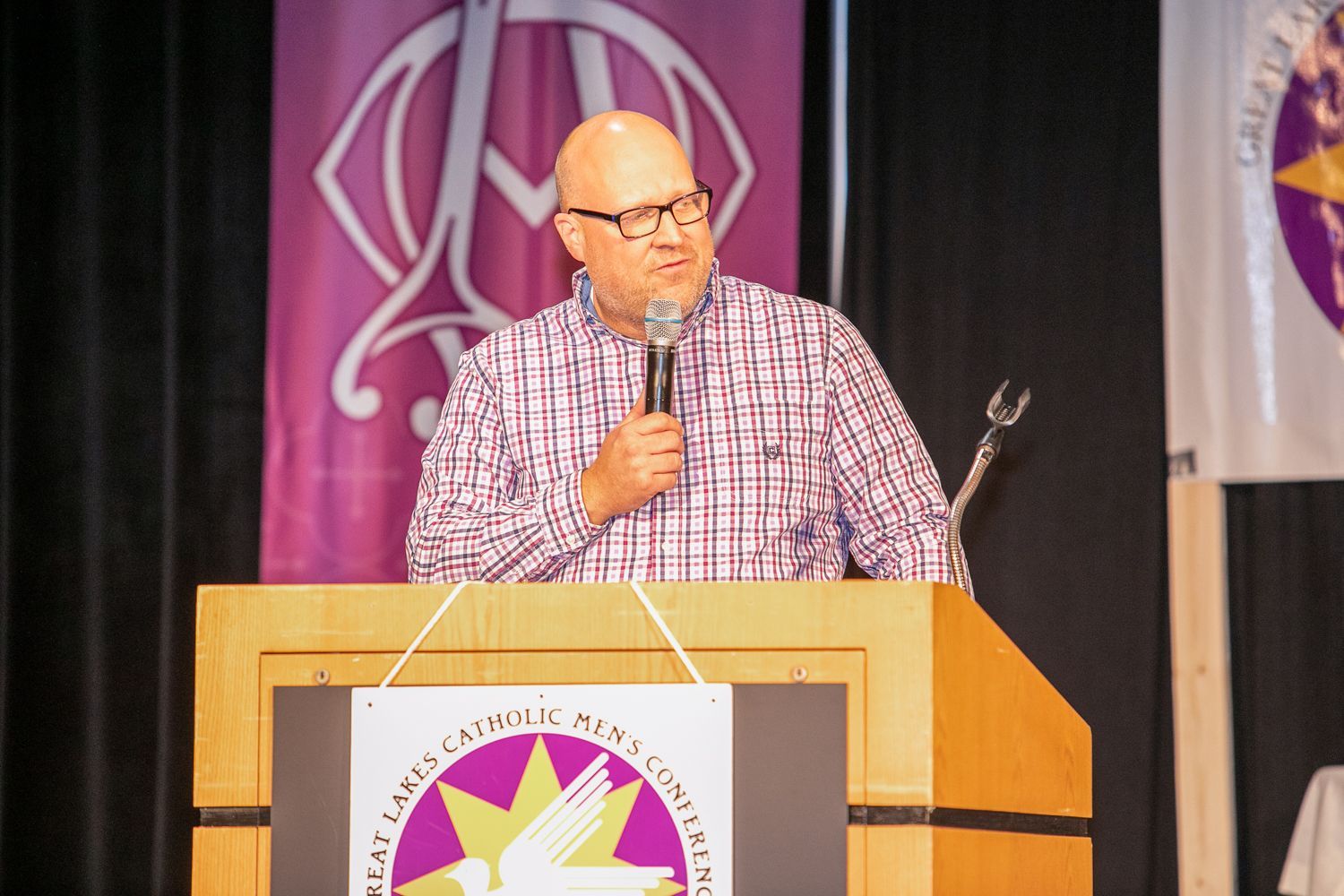 Mitch Striedl speaking at a podium on stage, wearing a plaid shirt and holding a microphone.