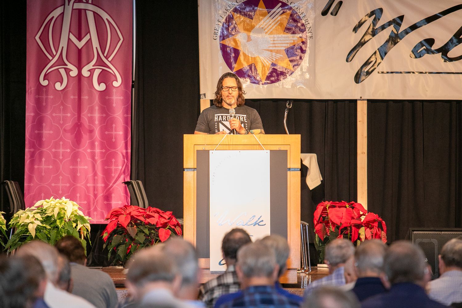 Keith Nester at podium addressing an audience in a decorated auditorium with banners and poinsettias.