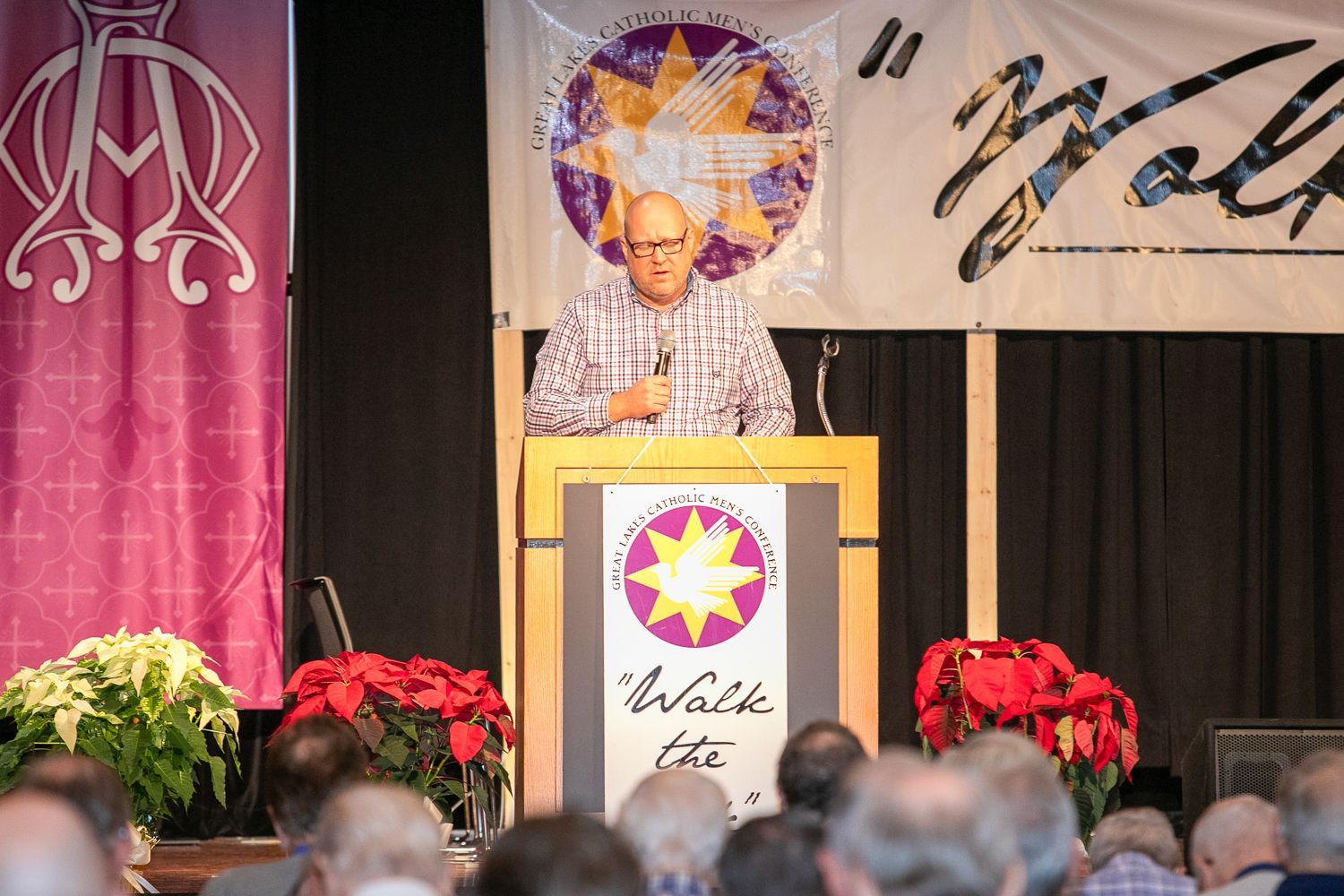 Mitch Striedl at podium on stage with floral arrangements and banners behind him