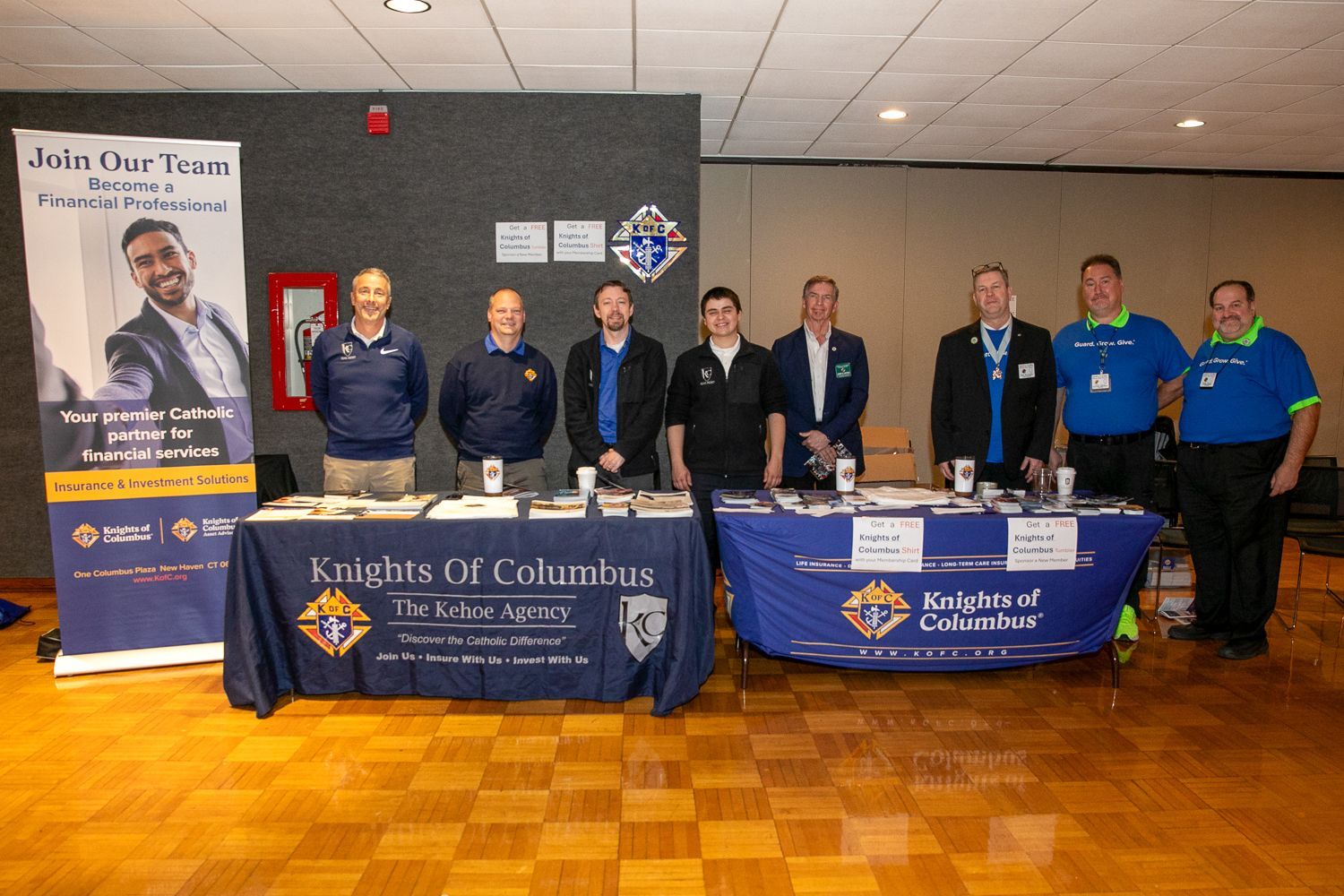 Group of eight people standing behind two blue event tables in a hall with a banner display.