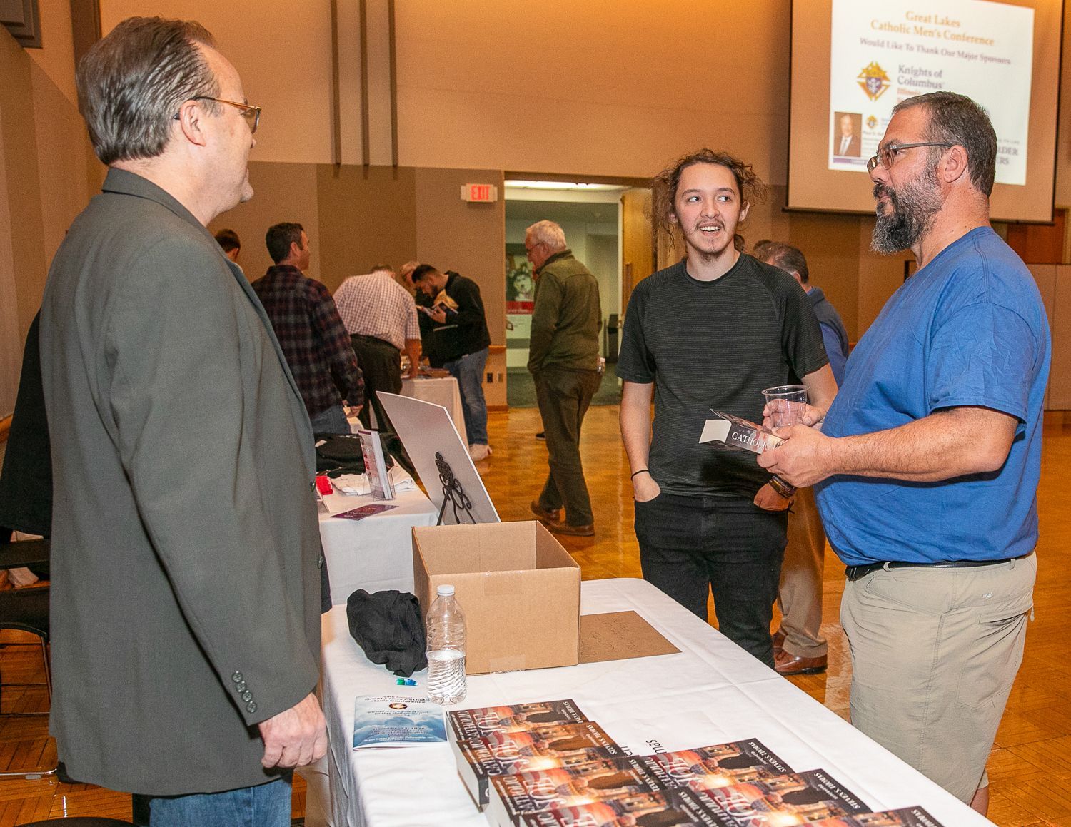 Three people chat at a trade show booth with brochures and boxes on a table.