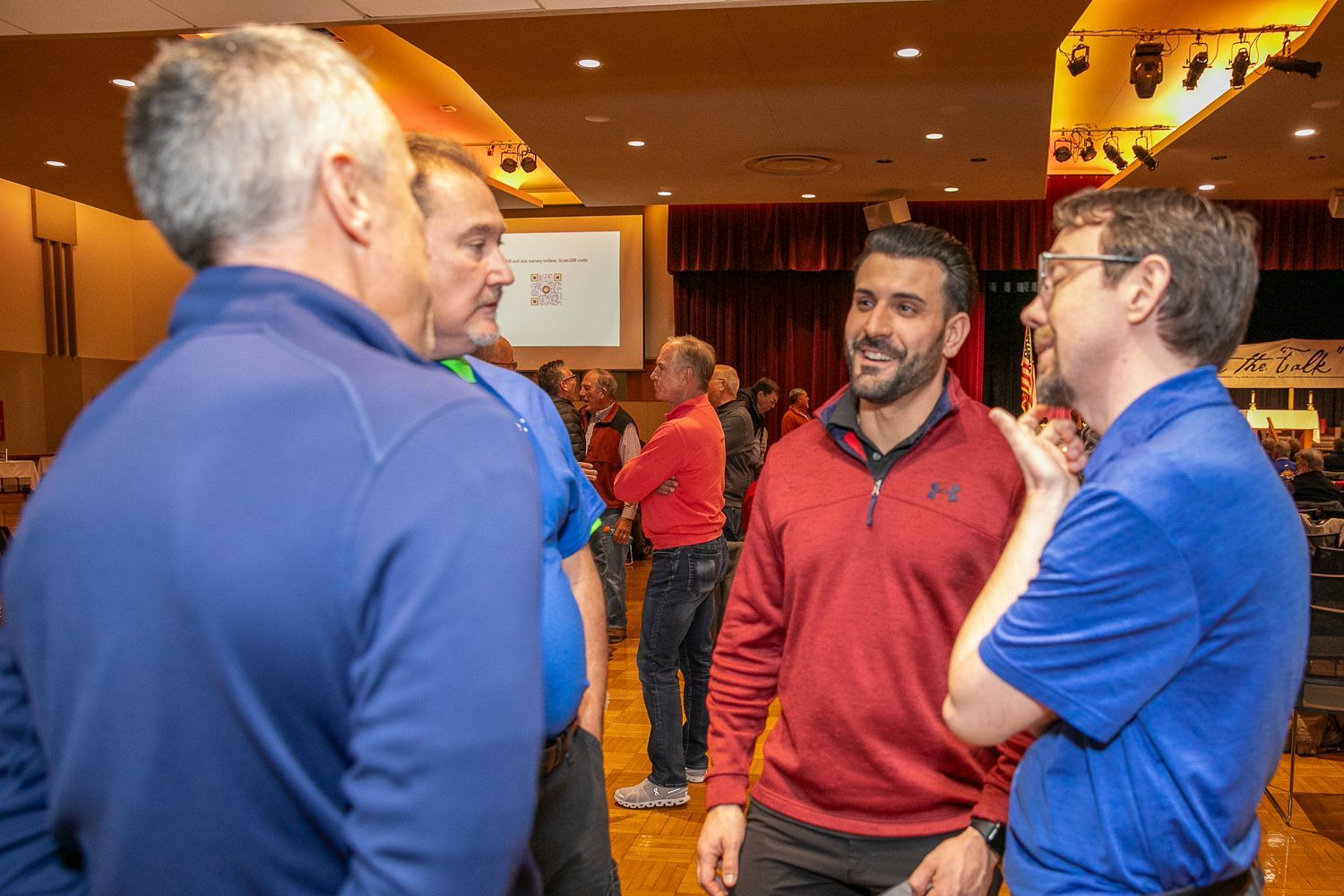 People chatting in a warm-lit indoor lobby, two men in blue shirts facing a man in a red pullover.