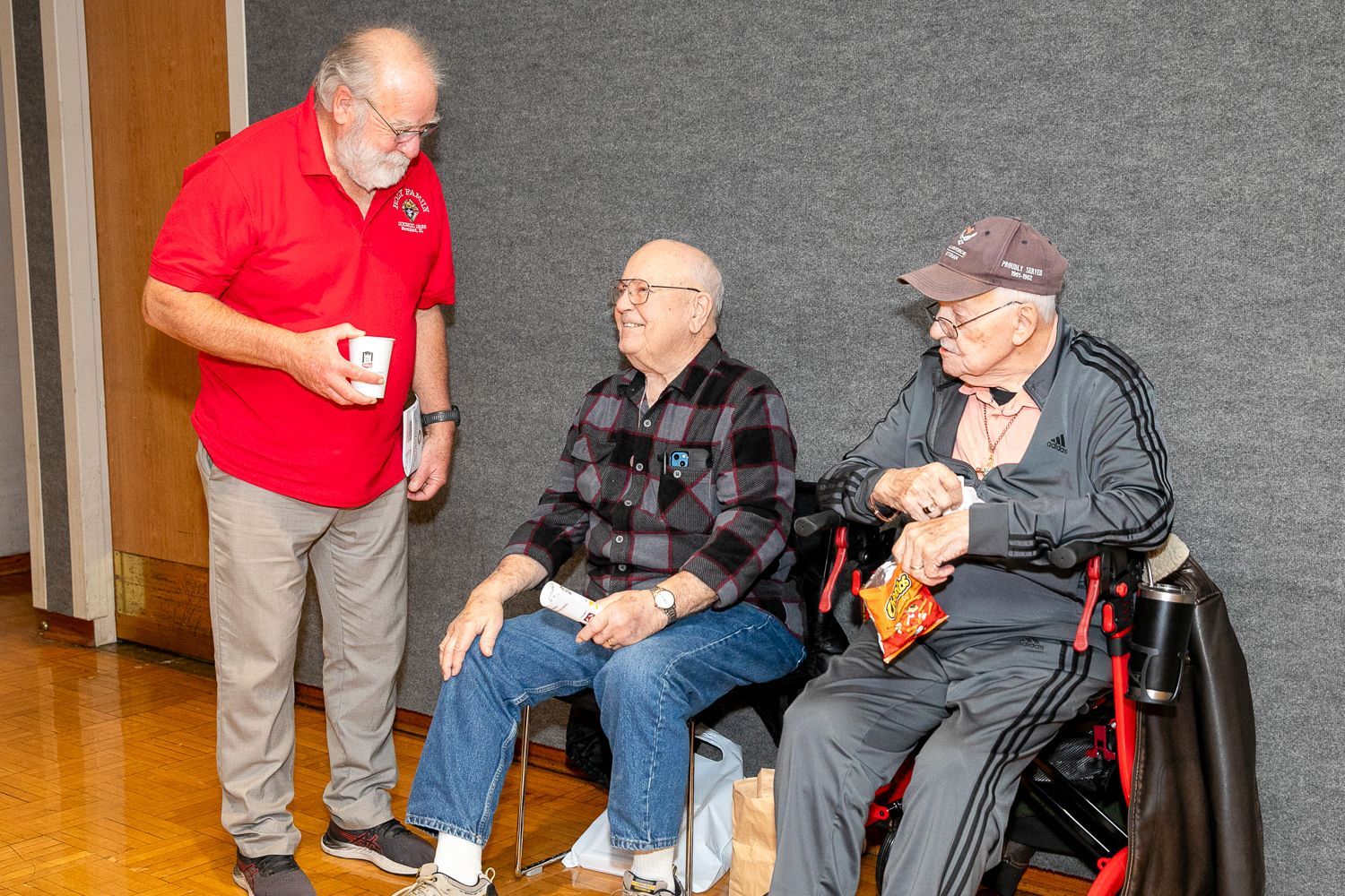 Three men chatting indoors; one stands in a red shirt while two sit, one in a wheelchair.