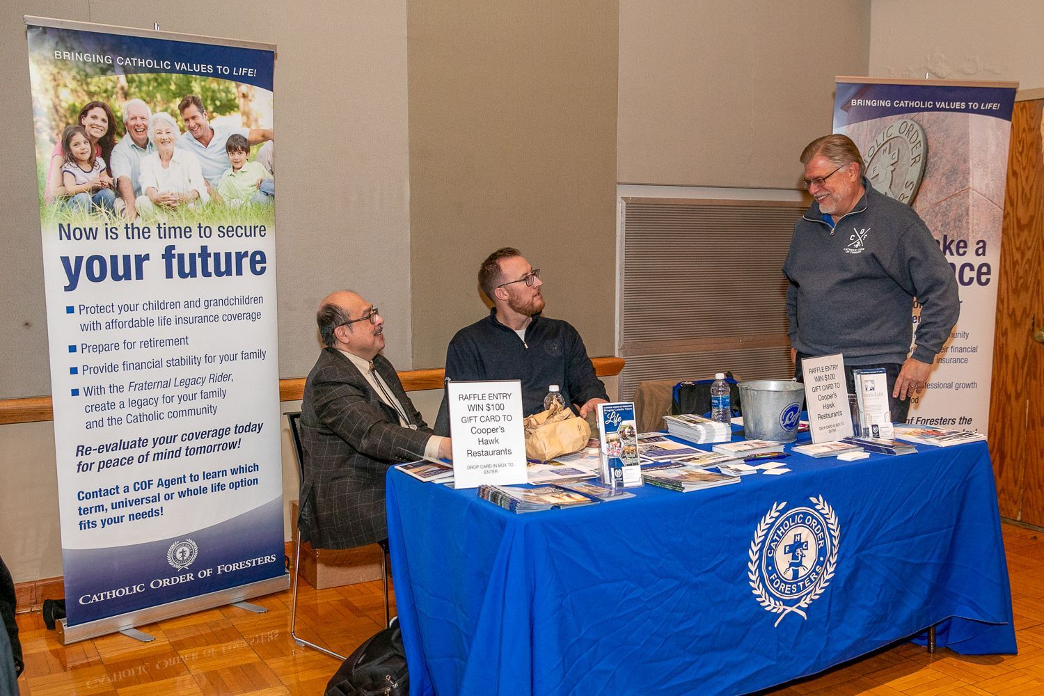 Three people at a blue-covered booth with banners and brochures at an indoor event