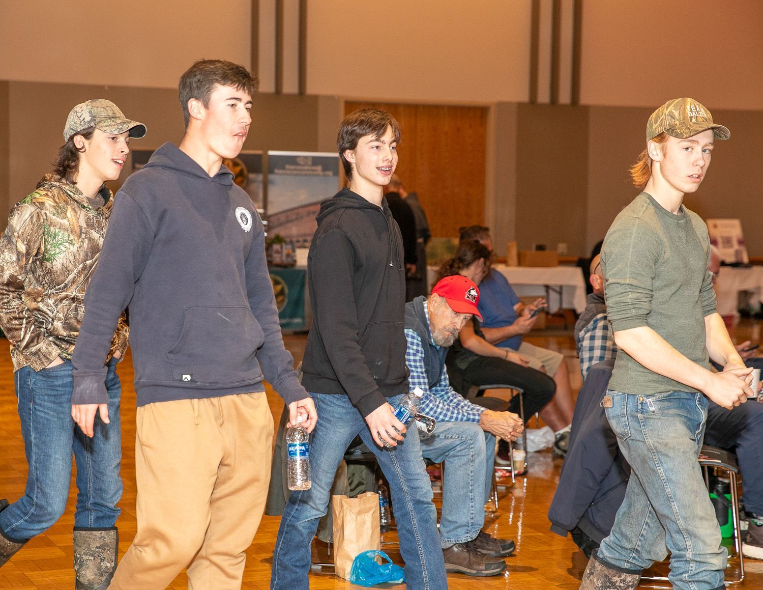 People walking indoors in a gym, with one person in a navy sweatshirt and tan pants in front.