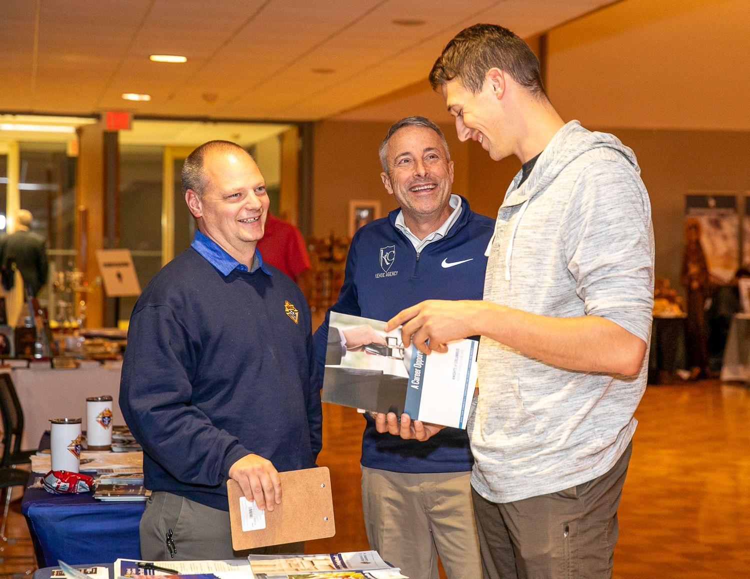 Three men at an indoor event examining a bag and papers at a table with displays behind them