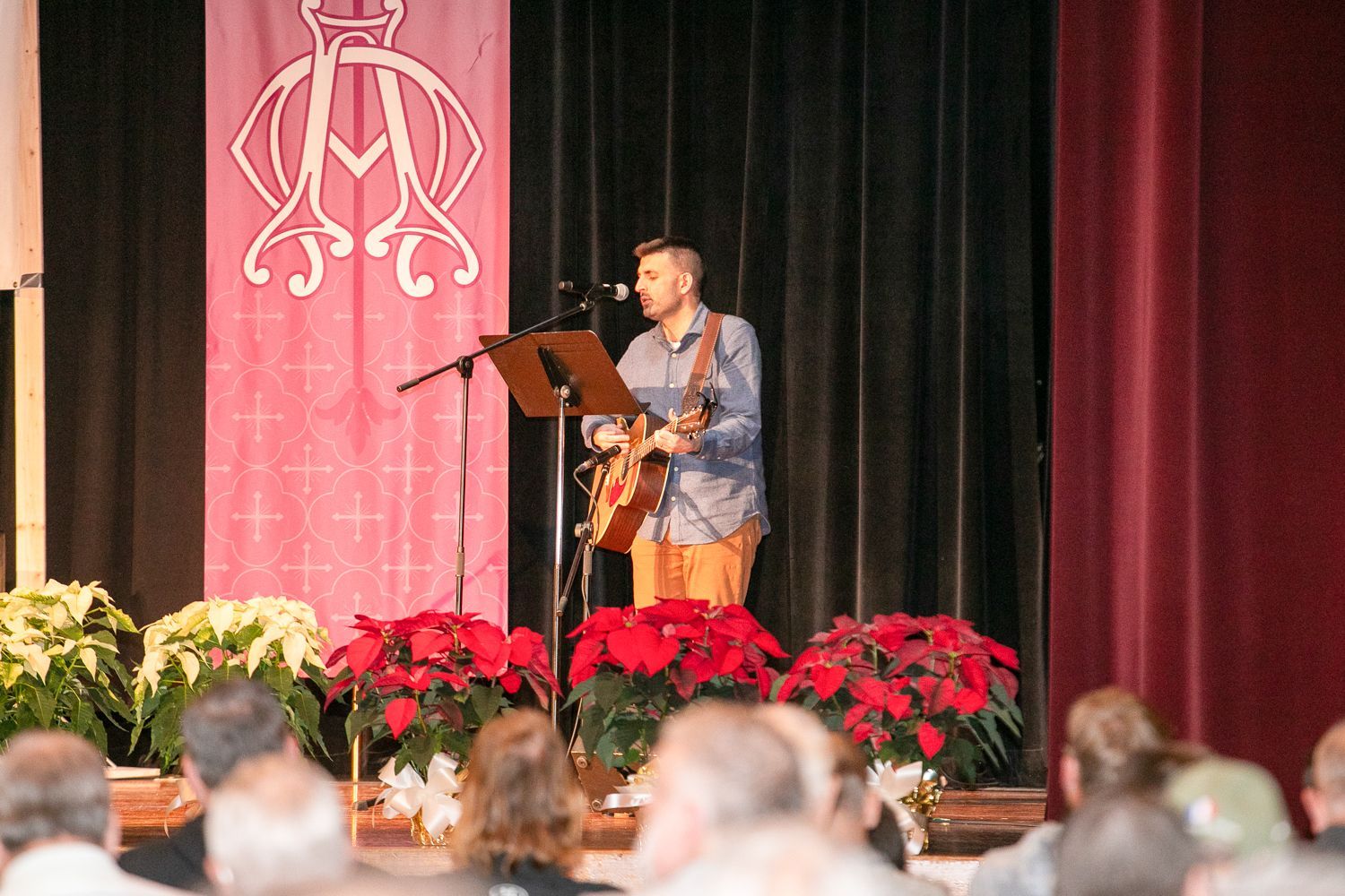 Musician singing and playing guitar on stage beside a microphone stand and red floral arrangements