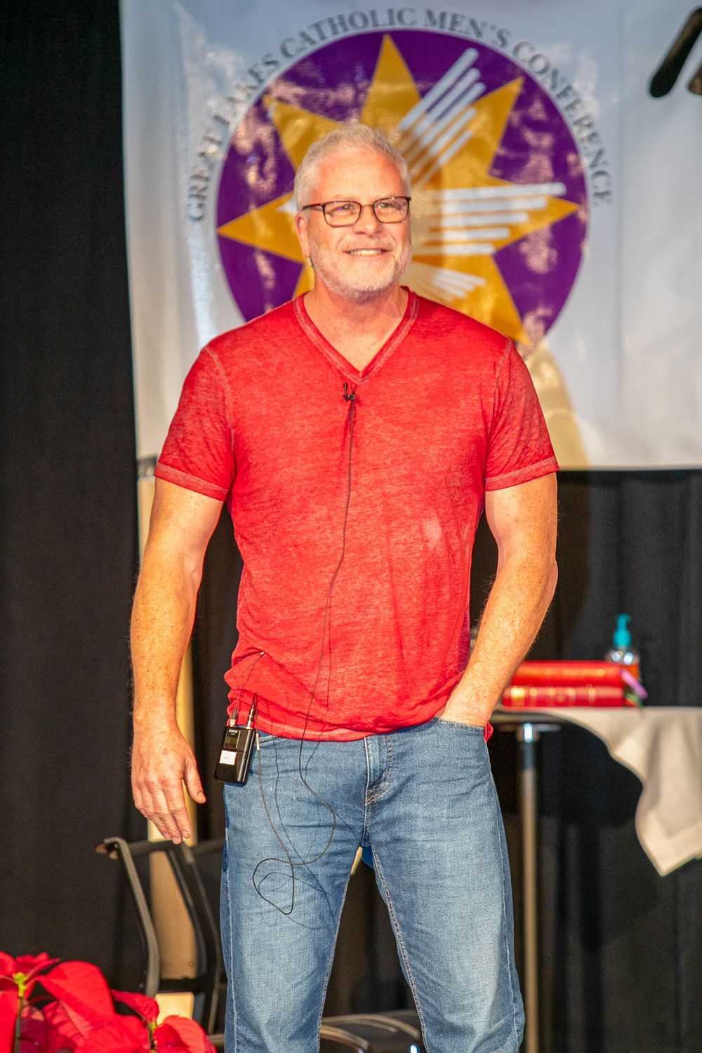 Sean Forrest in a red shirt and jeans standing on stage in front of a banner with a star emblem.